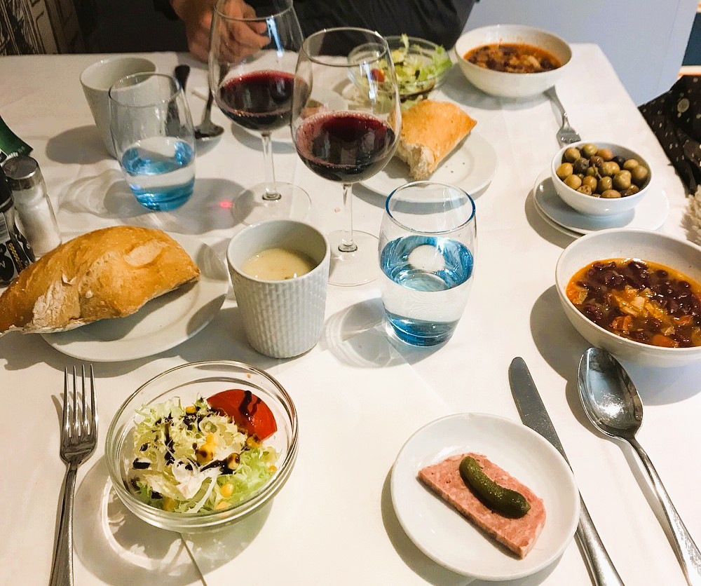 Un restaurant clàssic al barri antic de la capital Mínim's: una cuina Andorrana i de mercat en un entorn encantador al al centre del Centre Històric.

Hi ha dies en què ja saps que aniràs a un lloc i, només de pensar-hi, se’t dibuixa un somriure. Això és el que em passa amb un petit restaurant d’Andorra la Vella que m’ha robat el cor: per la seva cuina honesta i per la parella que hi ha al darrere, que porten el negoci amb ofici i molta ànima. Són d’aquells amfitrions que et truquen pel nom, t’escolten i aconsegueixen que cada visita sigui una mica especial.

L’espai té aquella personalitat dels locals de tota la vida: un baix d’una casa antiga aprofitat com abans es feia a l’Andorra més autèntica, amb parets que semblen guardar històries i un menjador dividit en racons que conviden a la conversa. Només travessar la porta, la vista se’n va inevitablement cap a la cuina oberta i als fogons, on alguna cassola deixa anar un xup-xup prometedor i alguna paella deixa ballar bolets a la paella. Mentre et saluden, t’acompanyen a una taula propera a l’entrada, i de seguida notes aquella calidesa de casa de poble: llum amable, soroll suau de coberteria i converses en segon pla.

Entre setmana hi fan un menú pensat amb cap i cor, amb platets que canvien segons mercat i temporada, ideal per descobrir sabors sense haver de triar massa. En cap de setmana, però, la carta es desplega amb plats per compartir i elaboracions que demanen temps i paciència: un carpaccio de vedella ben fi amb oli aromatitzat, verdures de temporada saltejades amb marisc, un bon peix al forn amb patata i ceba, talls de carn servits amb patates fregides de veritat i cebes tendres marcades a la planxa. De tant en tant, hi apareix algun guisat de caça fet amb tres dies de feina, amb salses d’aquelles que demanen pa per sucar fins a l’última gota.

Abans que arribin els plats, et reben amb petits detalls: una mica de quiche tèbia, unes olives, embotit tallat ben fi i pa artesà amb crosta cruixent i molla tendra, d’aquells que només amb oli i sal ja serien un esmorzar complet. Quan comencen a desfilar els entrants, entens que aquí es cuina amb producte de veritat i amb respecte: verdures netes i tallades a mà, textures cuidades i combinacions que tenen sentit. Els principals mantenen el nivell, ja sigui amb un turbot que arriba al punt just o amb una peça de carn sucosa que es deixa tallar gairebé sense ganivet.

Les postres no són un tràmit, sinó una part més del ritual: un pastís de formatge per posar al mig de la taula i compartir, o un cafè irlandès servit com cal, amb les capes ben definides i aquell toc dolç que tanca el dinar amb elegància. Al final de l’àpat, la sensació és la d’haver estat a casa d’uns bons amics que cuinen de meravella: marxem satisfets, ben menjats, i amb la certesa que tornarem. És un d’aquells restaurants que no només alimenten, sinó que també cuiden, i això, avui, té un valor immens.